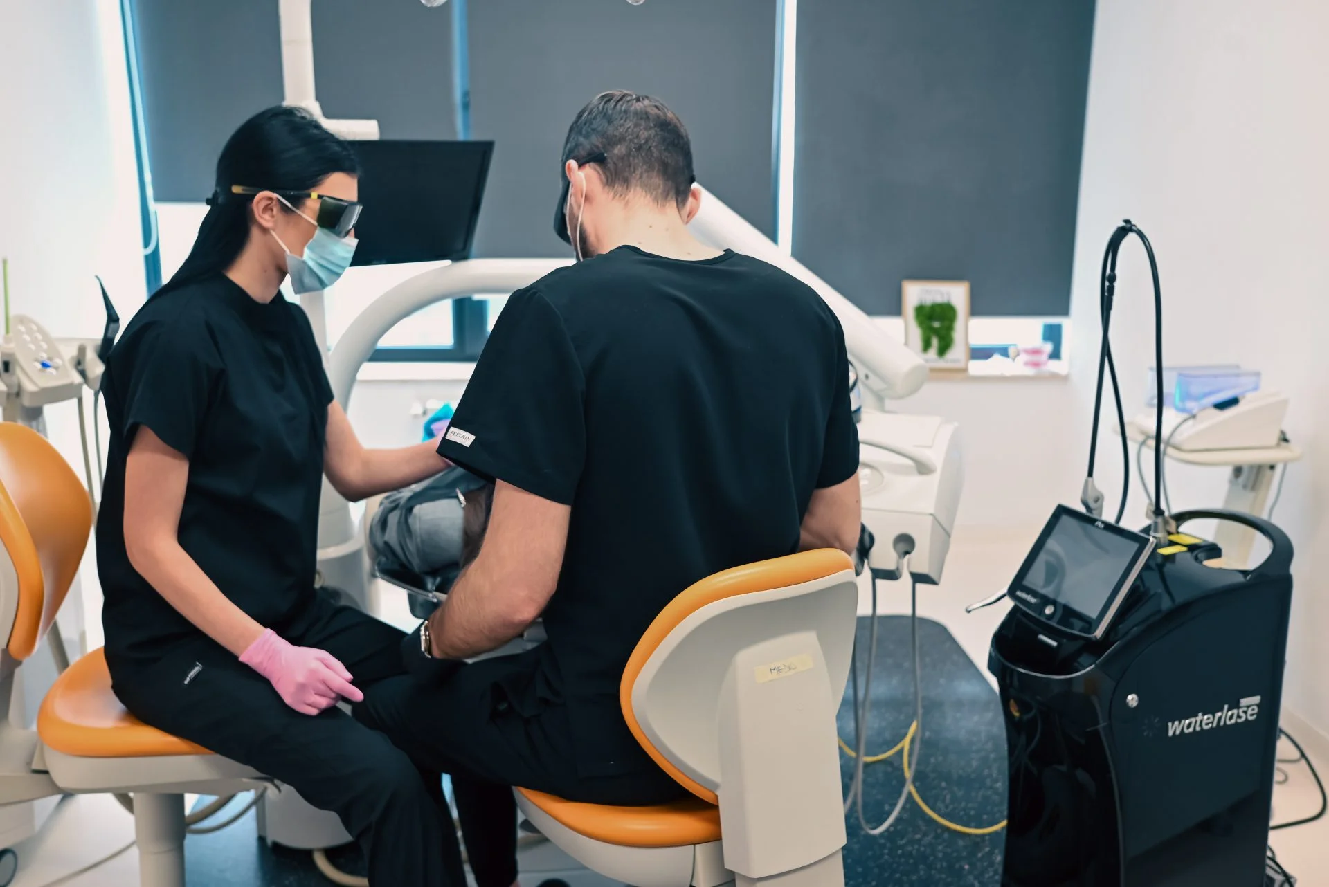 Asistenți în halate negre, echipament dentar, cabină estetică modernă.