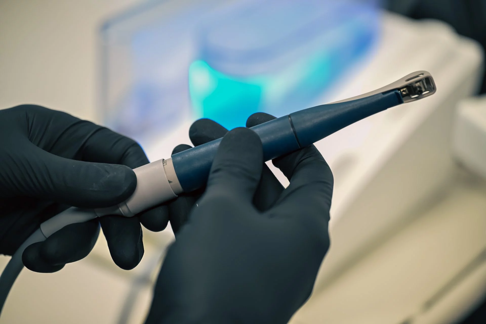 Două mâini cu mănuși negre țin un scanner intraoral albastru.