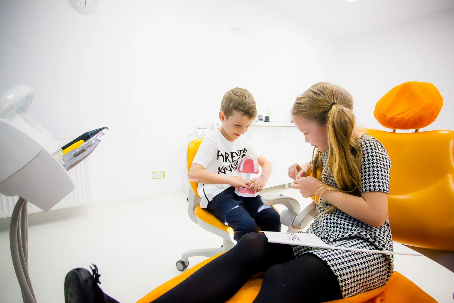 Copii examinând model dentar roz, așezați pe scaune portocalii.