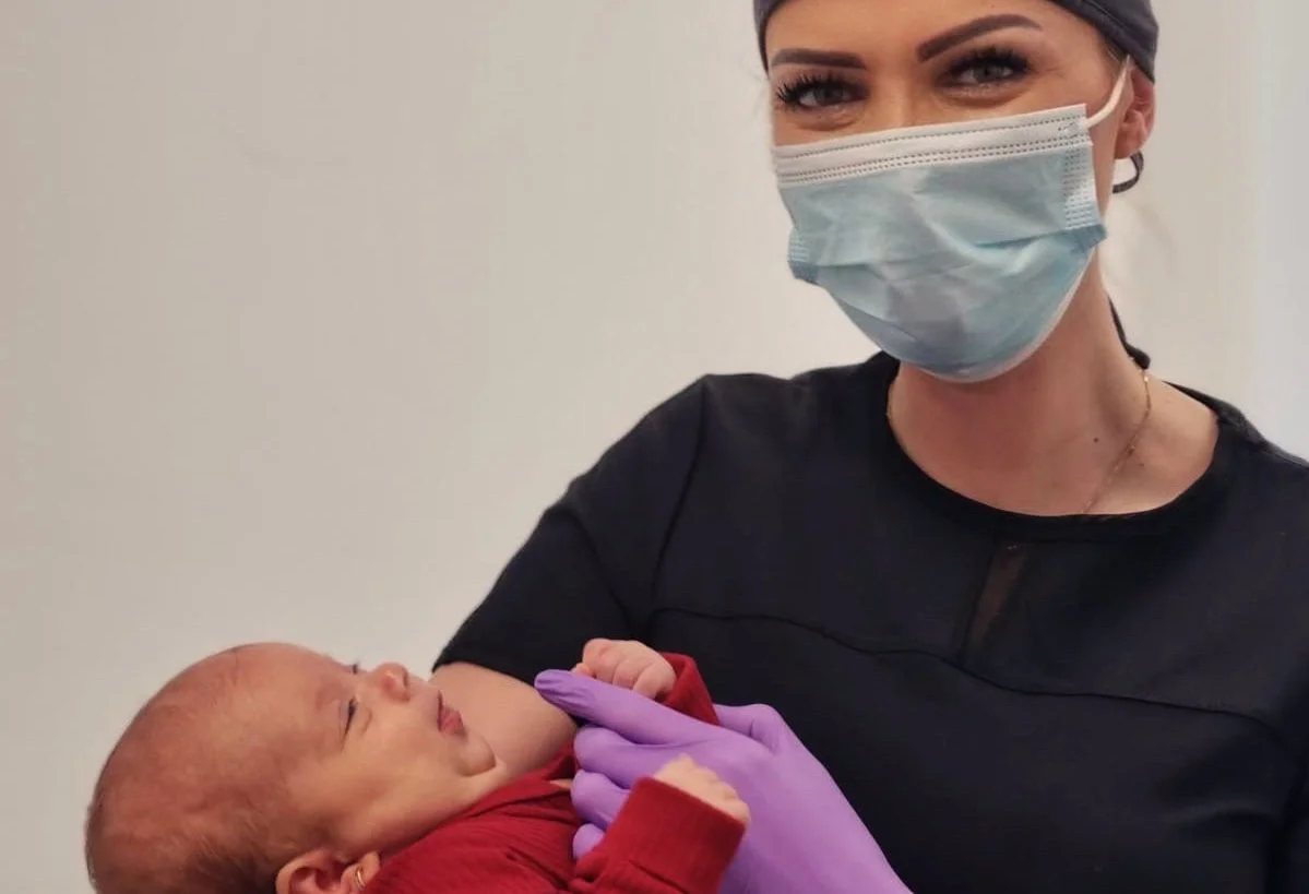 Persoană cu mască și mănuși mov ținând un bebeluș.